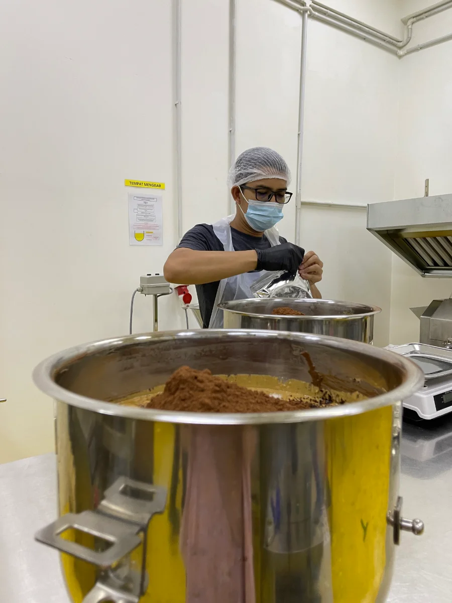 A food operator in hairnet, mask and gloves weighing spice powder into a large mixing pot in a production kitchen.